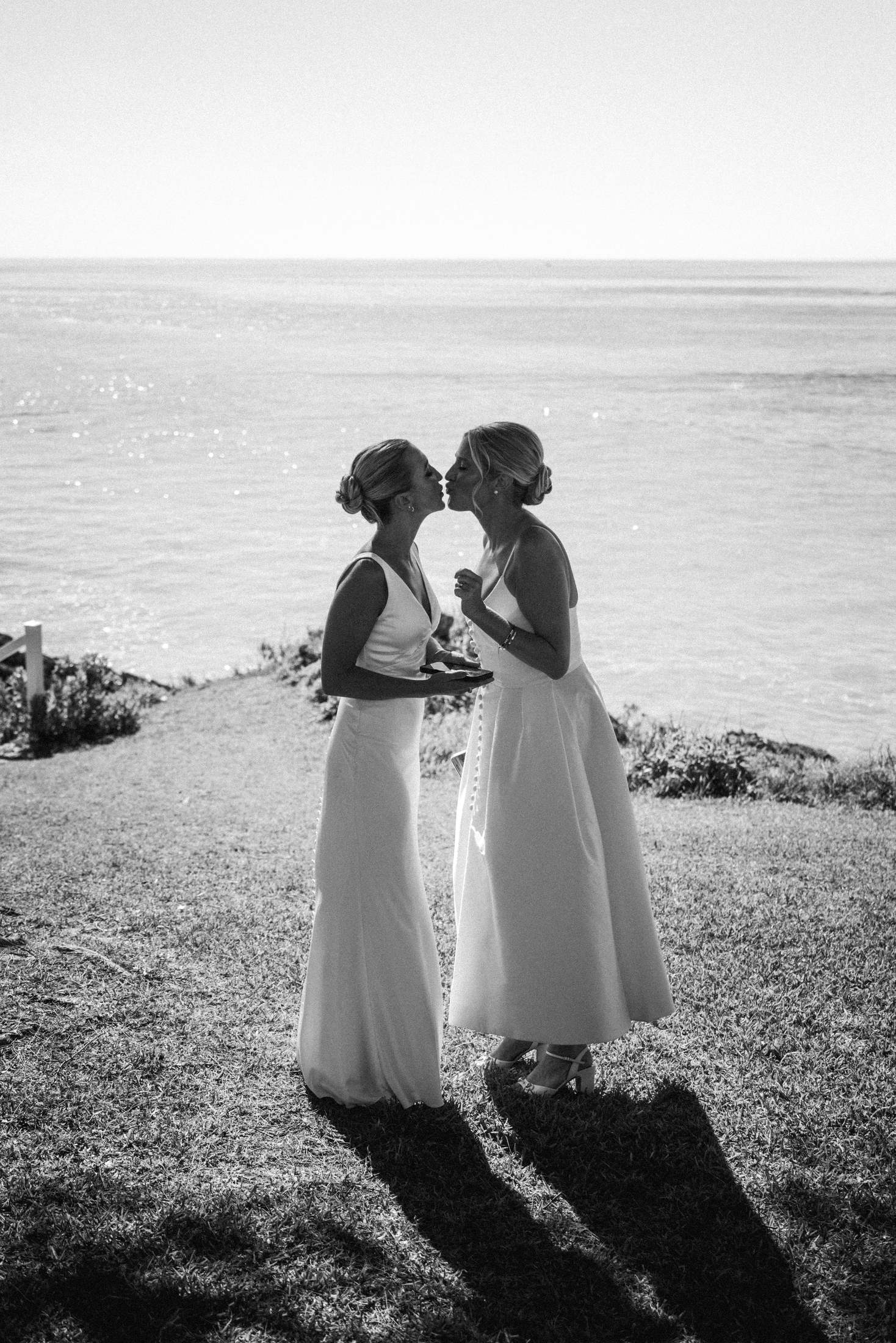 Black and white portrait of two brides forehead to forehead on a Bermuda cliff top with long evening shadows