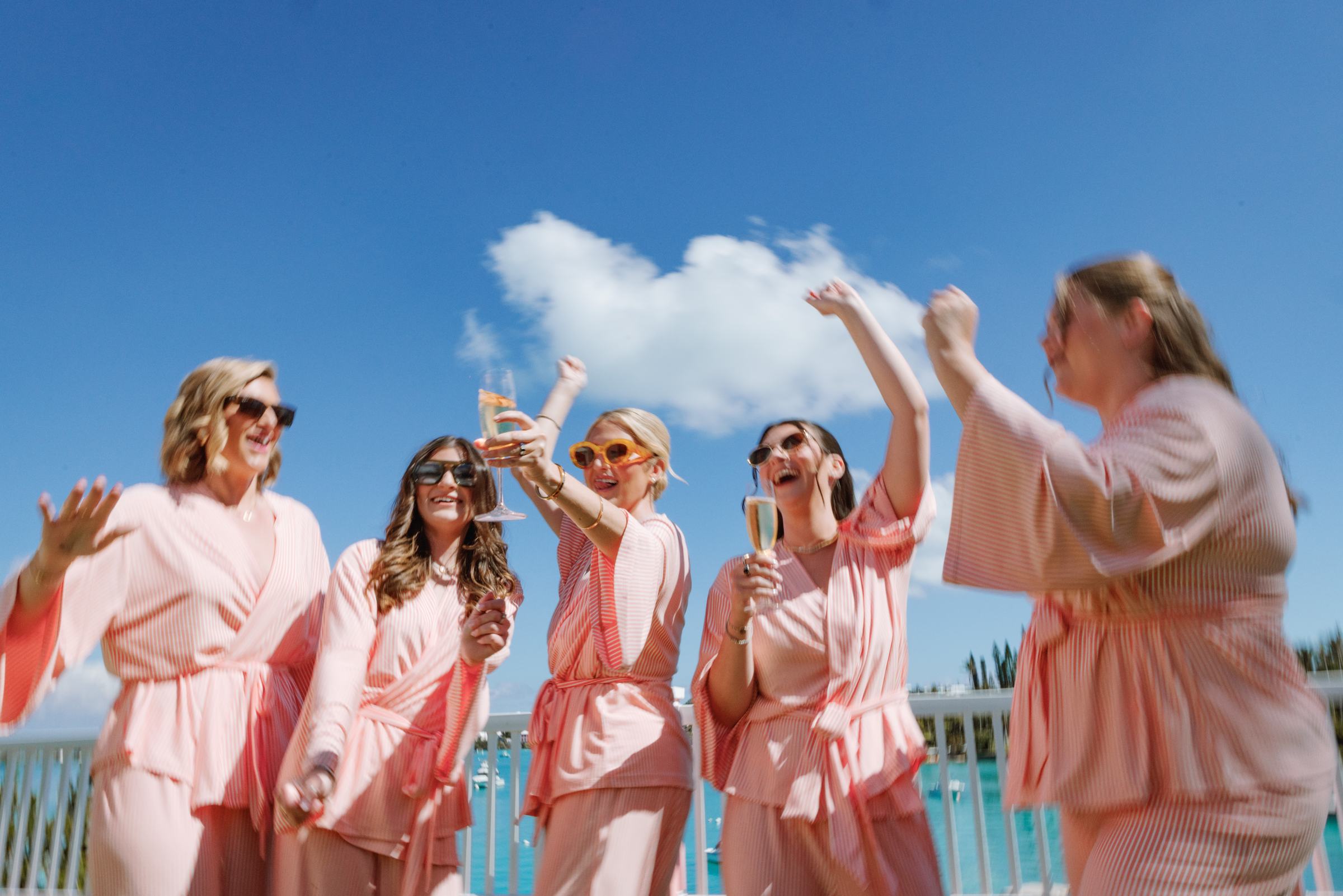 Bridesmaids in pink robes cheering with champagne under a blue Bermuda sky