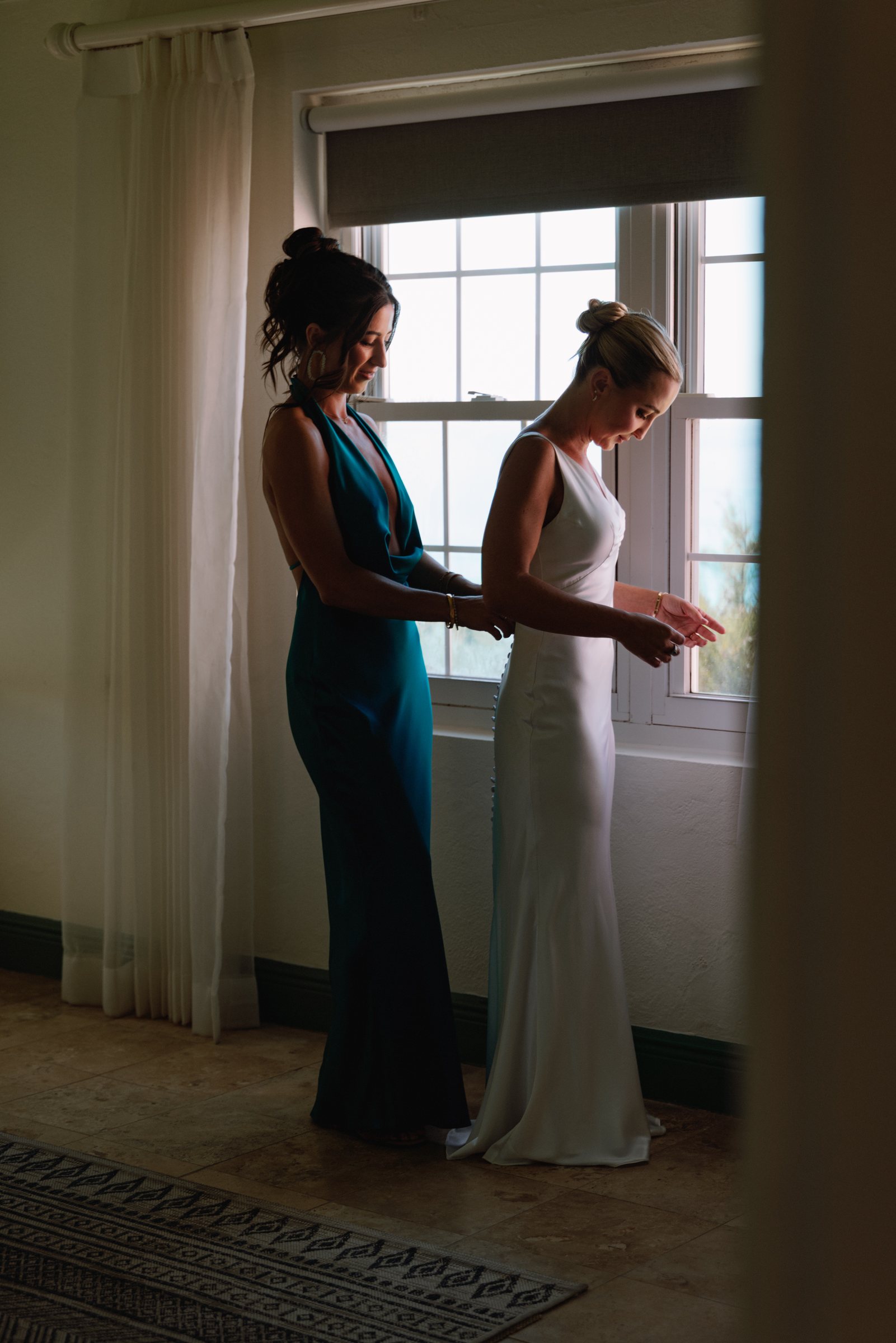 Bride being helped into her gown beside a bright window before her Bermuda wedding