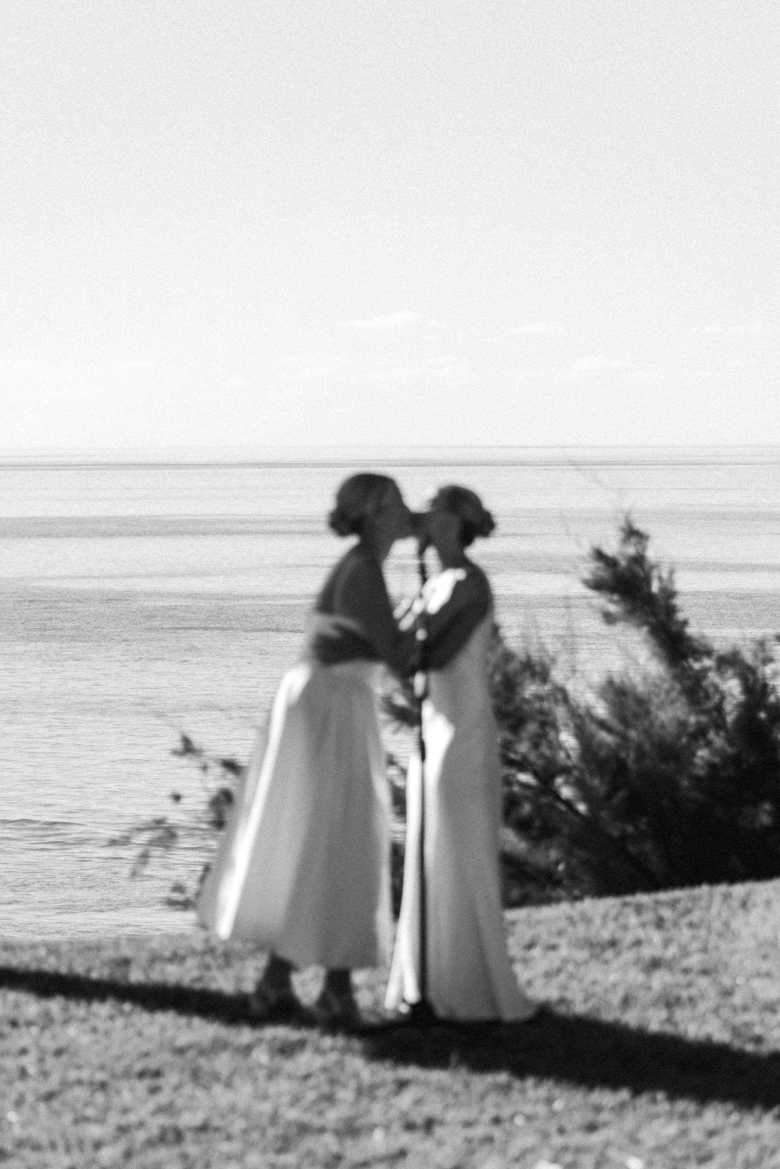 Black and white silhouette of two brides kissing on the cliff edge above the ocean