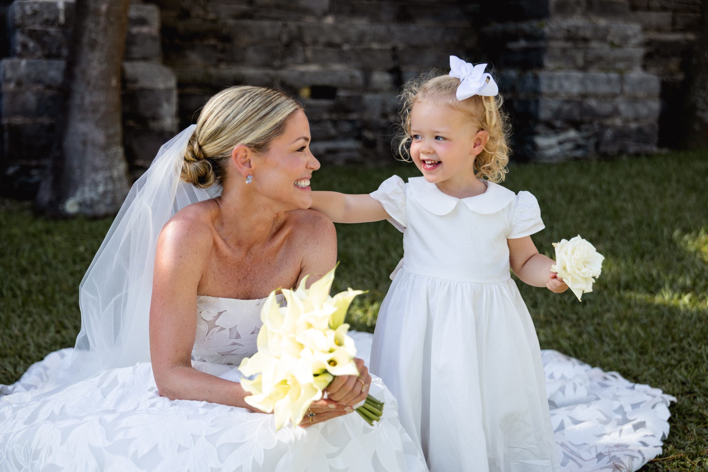 Bride sitting on the grass laughing with the flower girl at the Bermuda church ruins