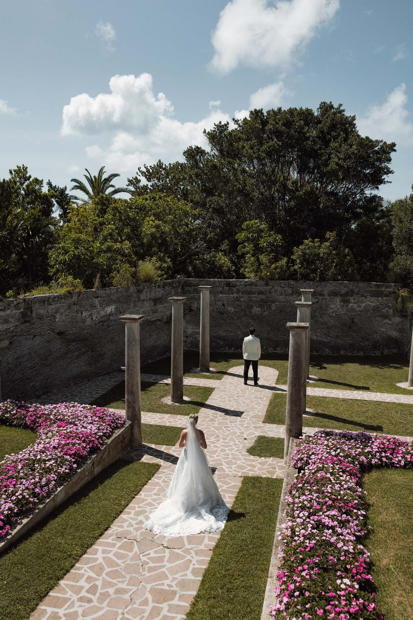 Bride walking the stone path between pink flower beds at the Bermuda church ruins
