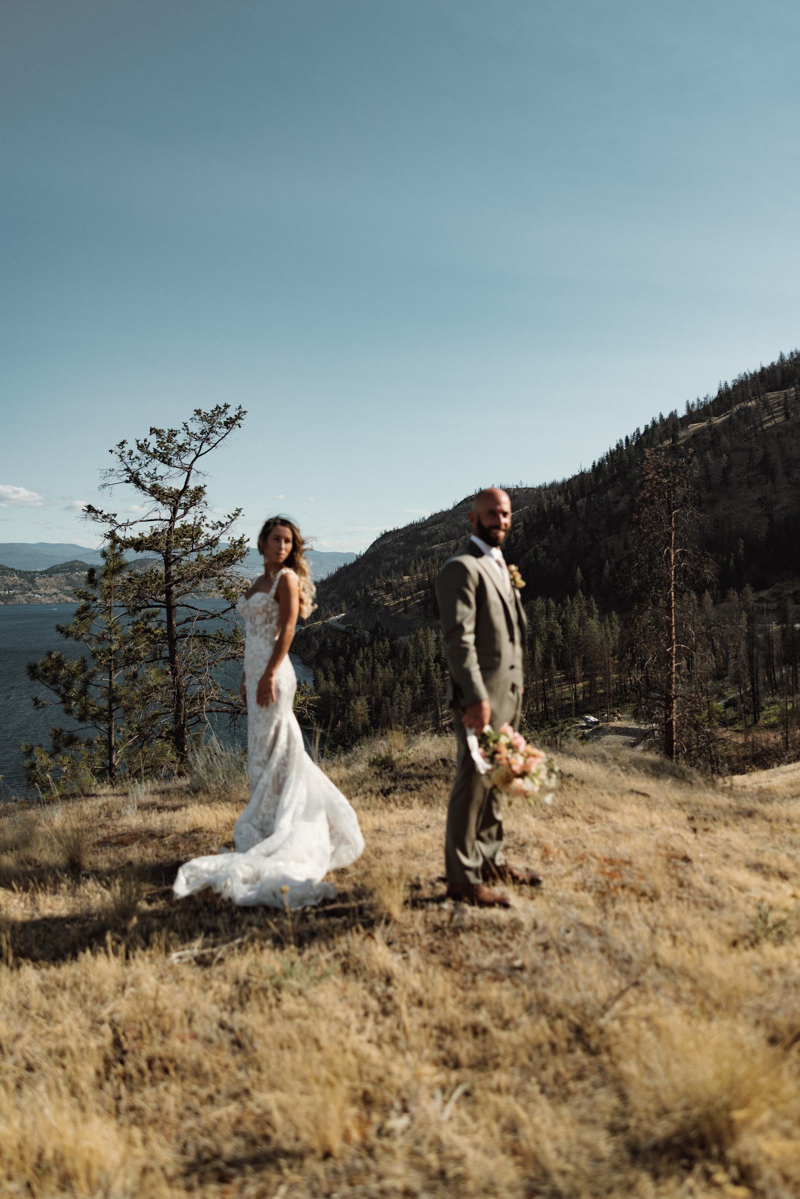 Bride on the golden hillside above the lake with the groom watching her