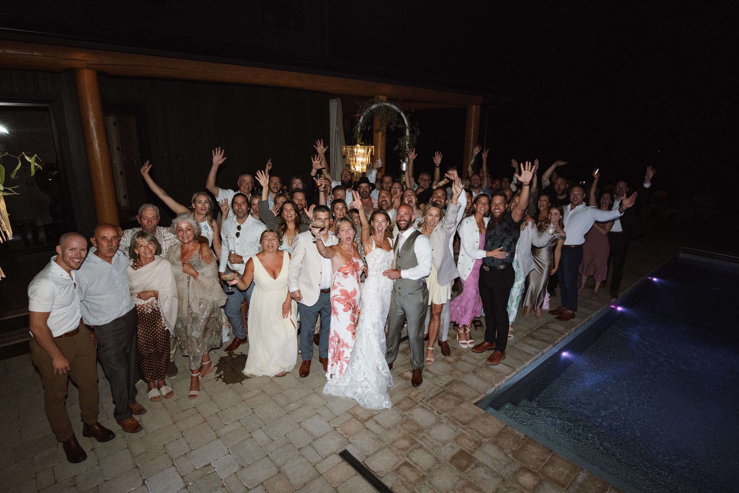 Wedding guests gathered by the pool at night with their arms raised