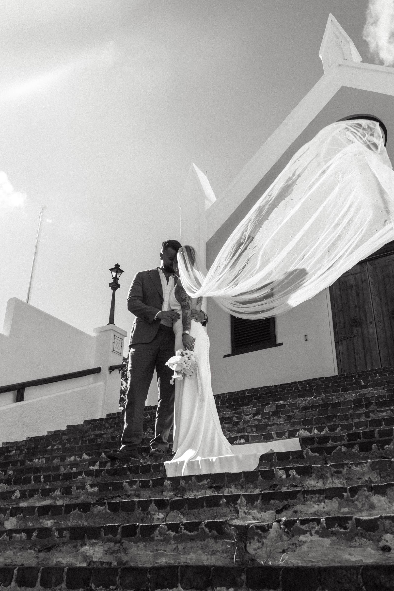 Black and white photograph of the couple on the church steps with the veil flying