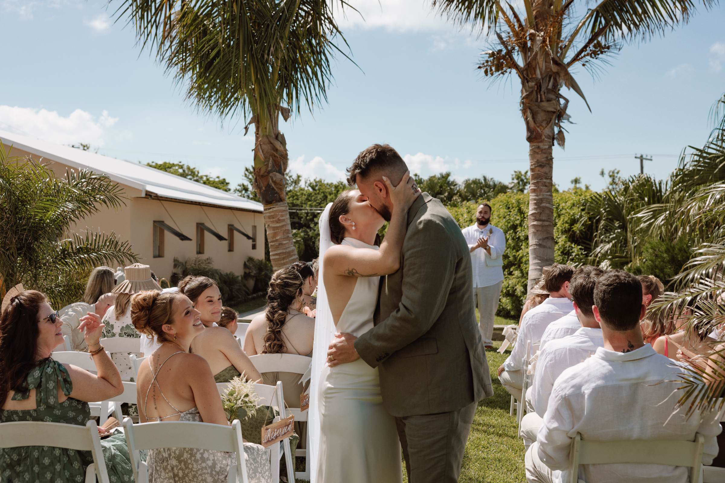 Bride and groom kissing under the palms as their guests look on