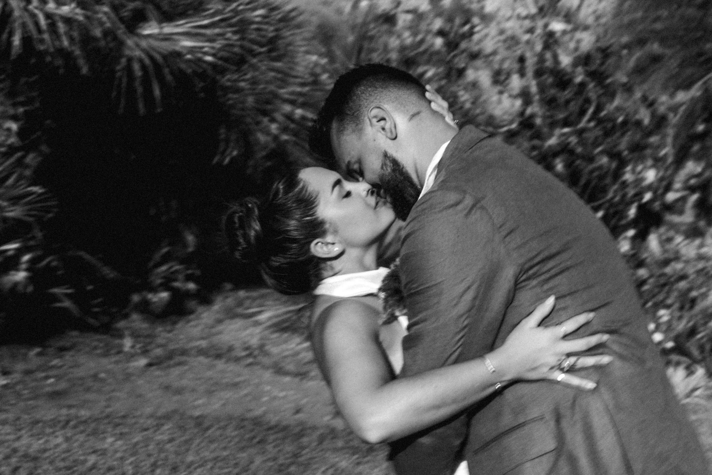 Black and white photograph of the couple kissing among tropical foliage
