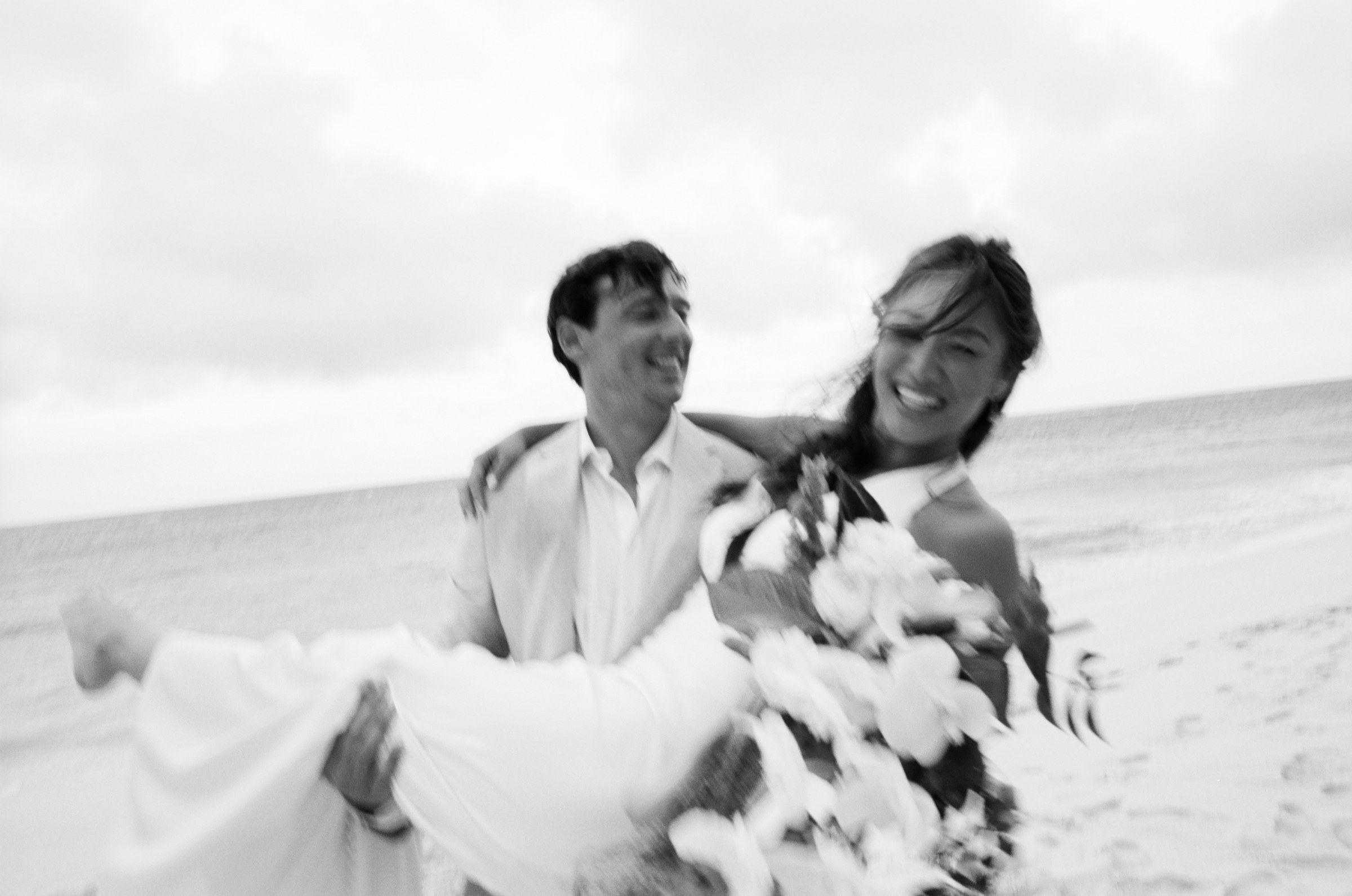 Black and white photograph of the groom carrying the bride across the sand, shot on film