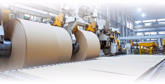 Large rolls of brown paper feeding into a machine in a spacious, well-lit industrial factory.