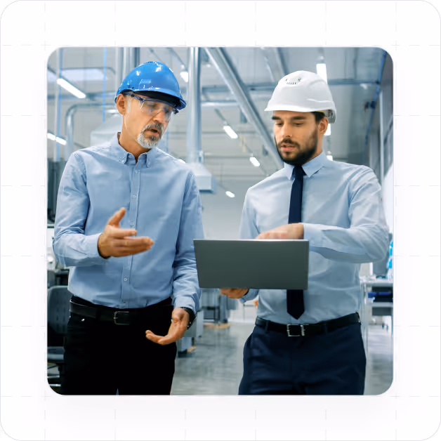 Two men wearing safety helmets and business attire discussing work while looking at a laptop in an industrial facility.