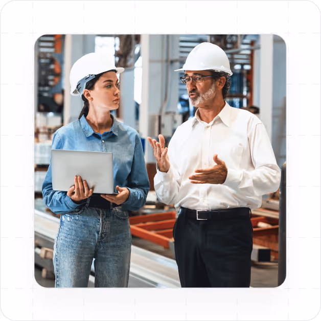 Two people in hard hats discussing work in an industrial setting, one holding a laptop.
