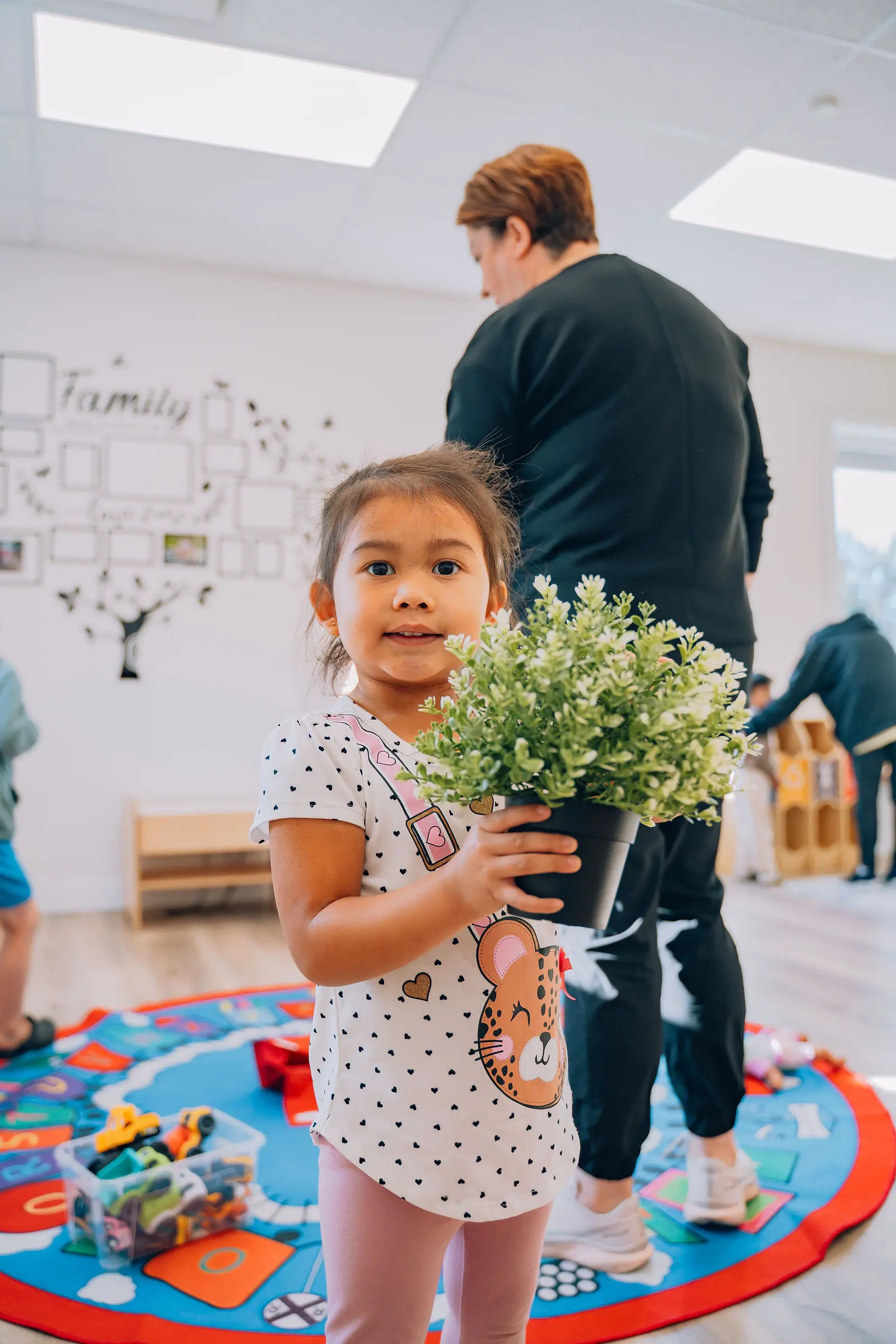 Little girl holding a potted green plant, standing on a colorful play mat in a classroom with adults and children in the background.