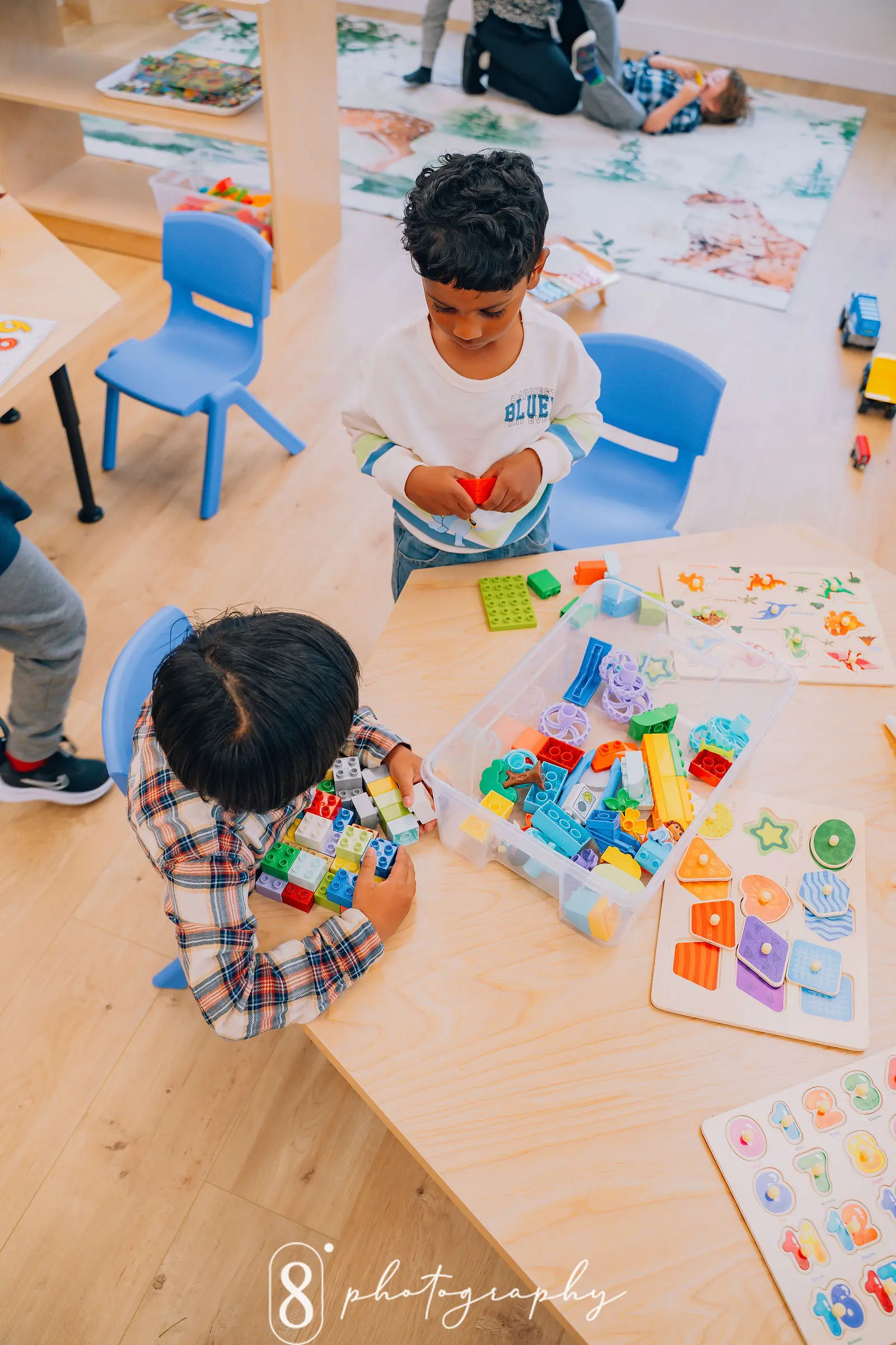 Two children playing with colorful building blocks and puzzles at a wooden table in a classroom.