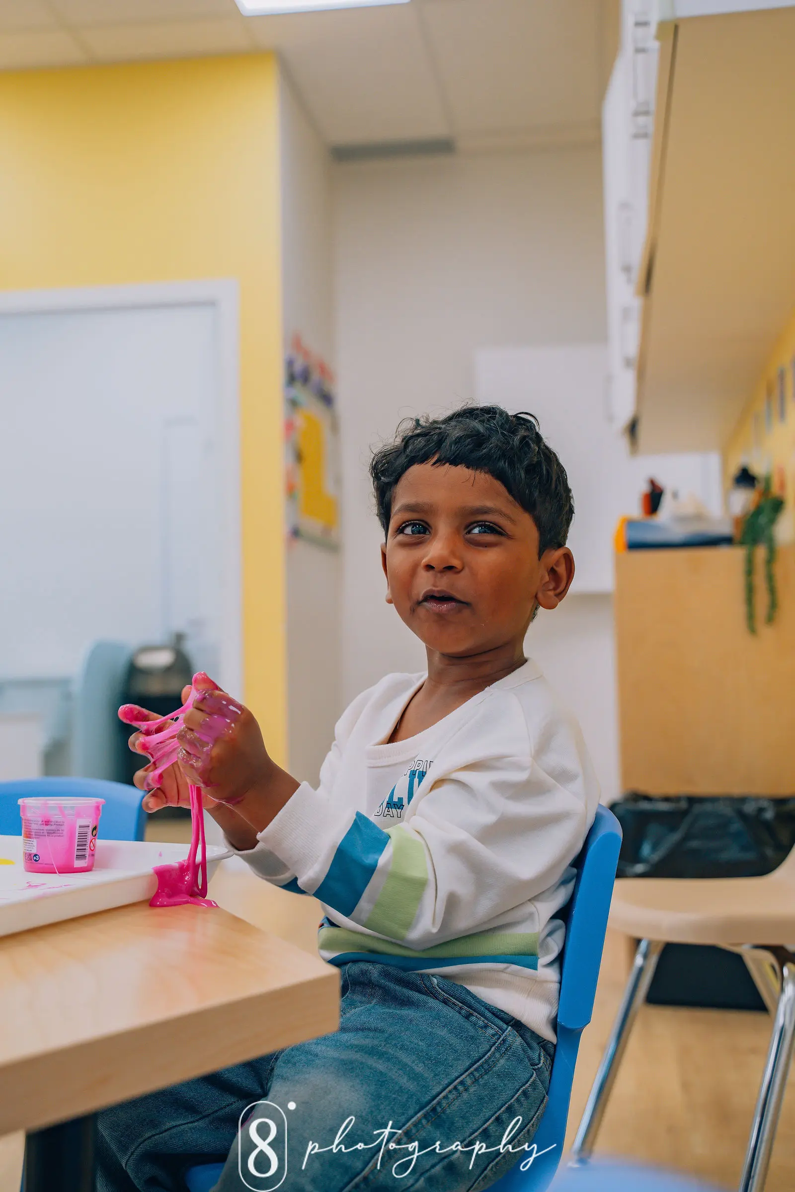 Young boy sitting on a blue chair playing with pink slime at a wooden table in a classroom.