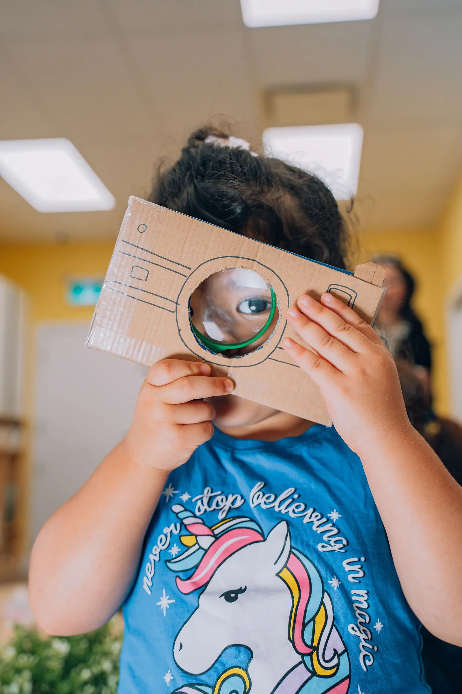 Child holding a cardboard cutout shaped like a camera up to their eye, wearing a blue shirt with a unicorn and the text 'never stop believing in magic'.