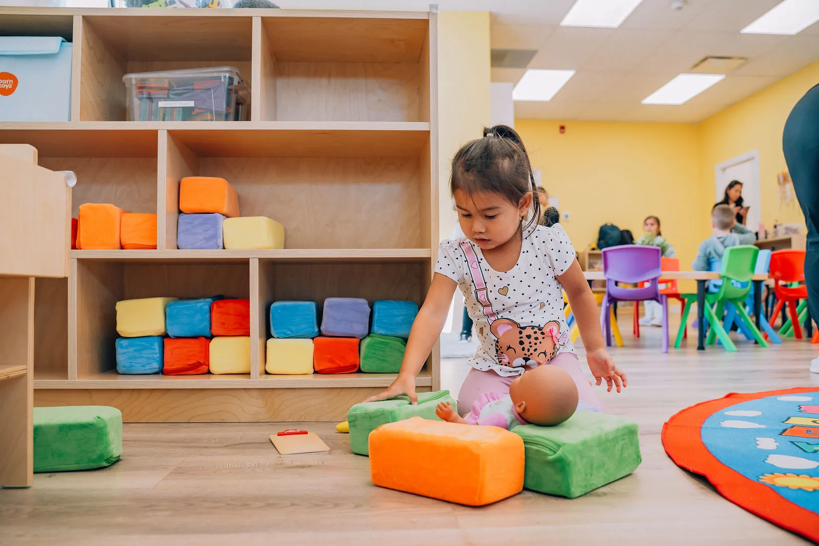 Young girl playing with a baby doll and colorful soft blocks on the floor in a brightly lit classroom with other children sitting at colorful chairs.