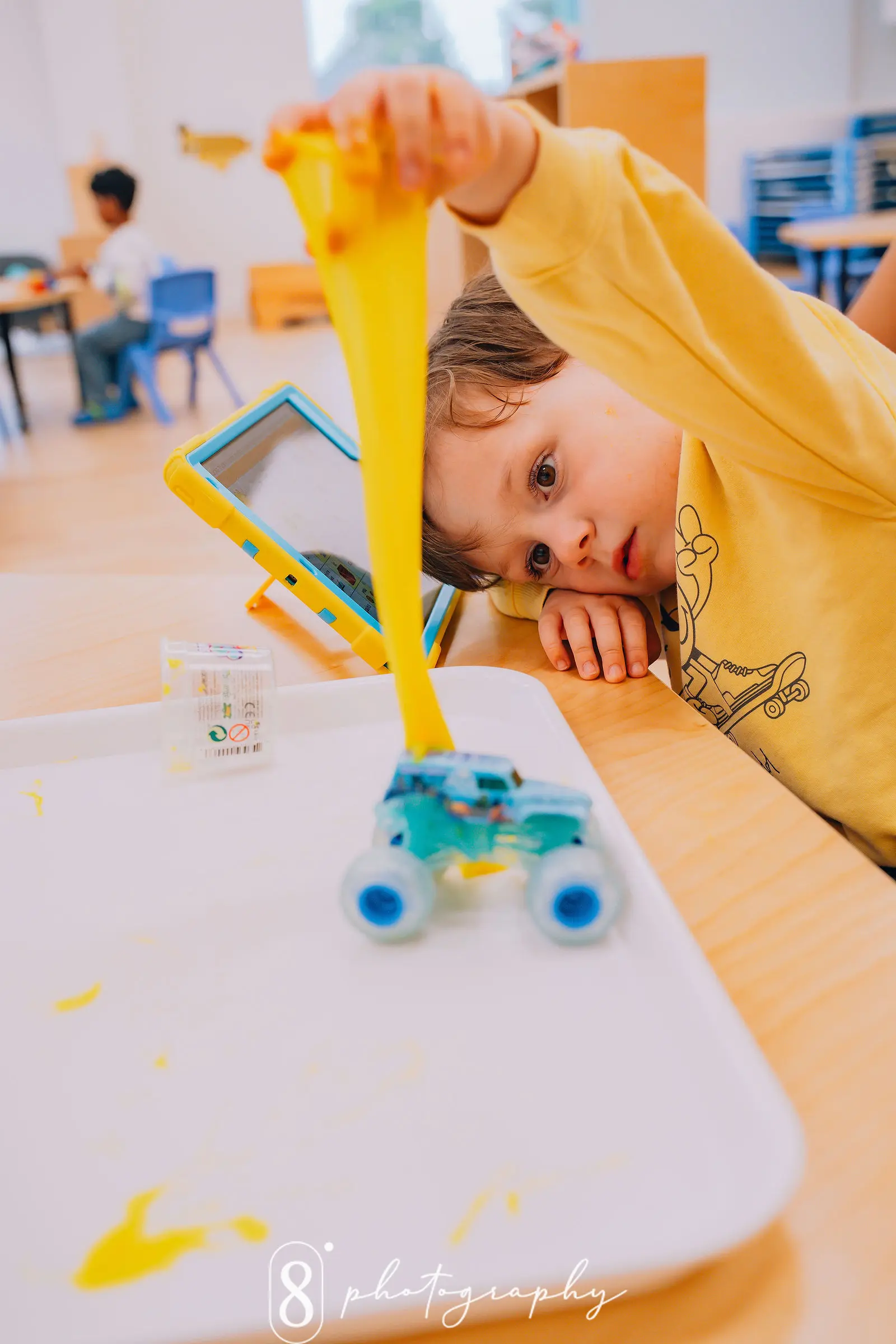 Young child in a yellow sweatshirt playing with yellow slime stretched above a blue toy car on a white tray.