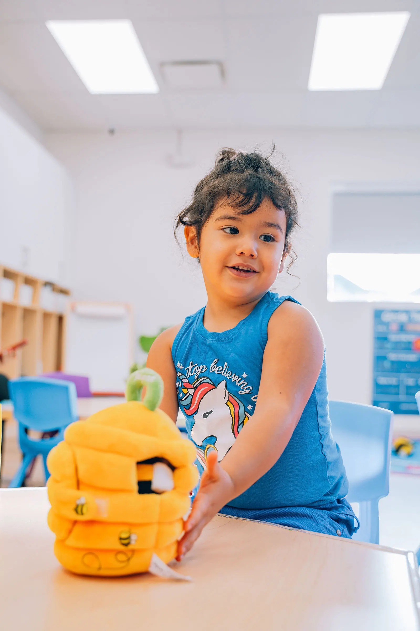 Young girl in a blue sleeveless shirt with a unicorn graphic playing with a yellow plush beehive toy at a classroom table.