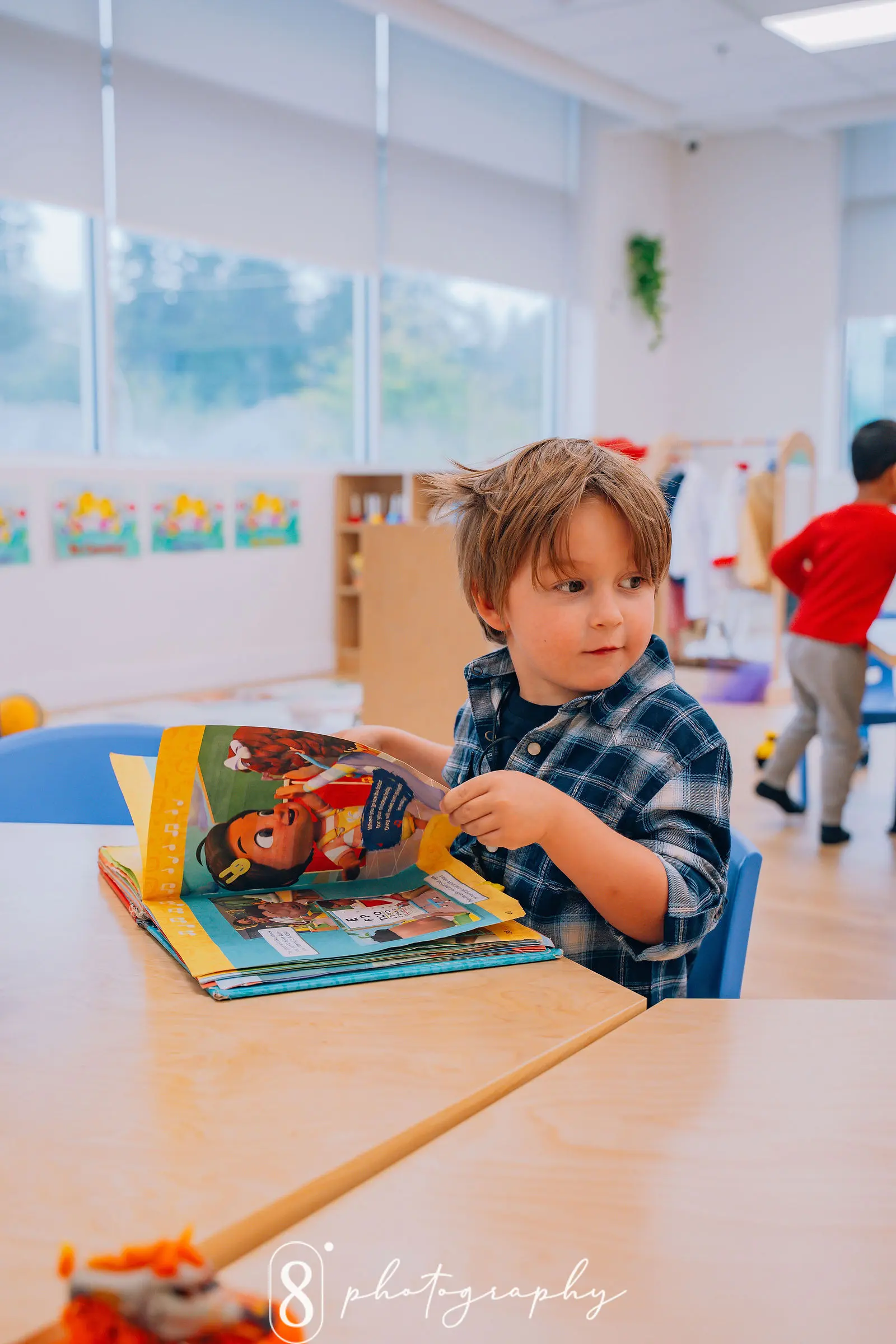 Young boy in a plaid shirt reading a colorful book while sitting at a classroom table.