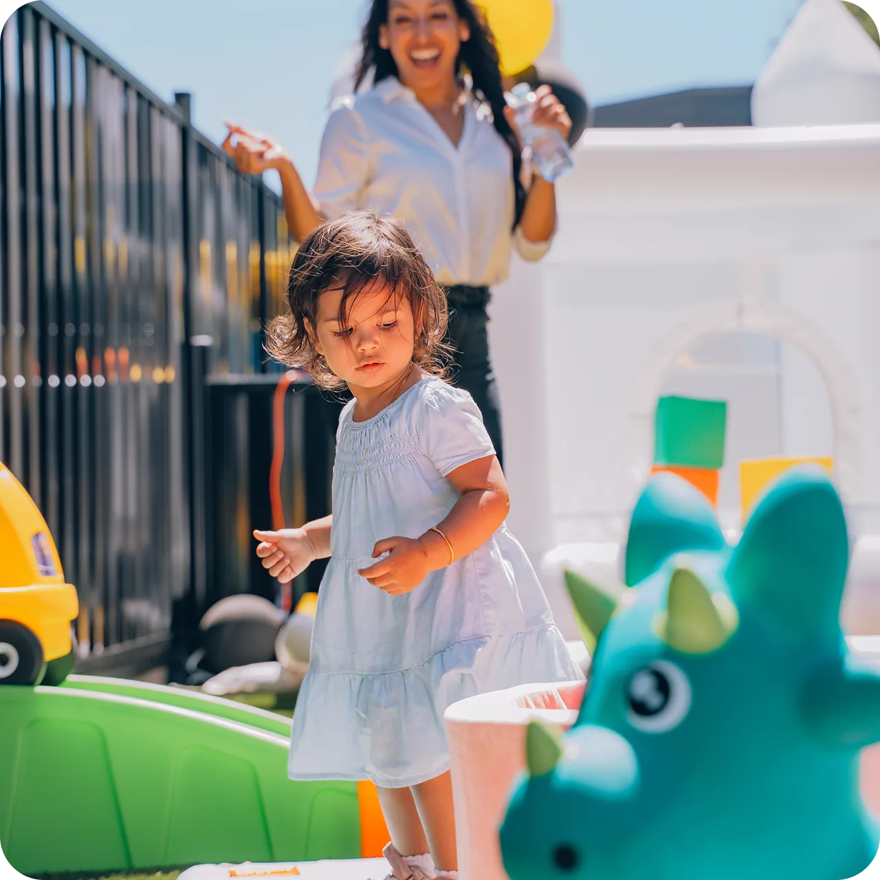 A toddler girl in a white dress playing outdoors with colorful toys while a smiling woman holds a water bottle in the background.