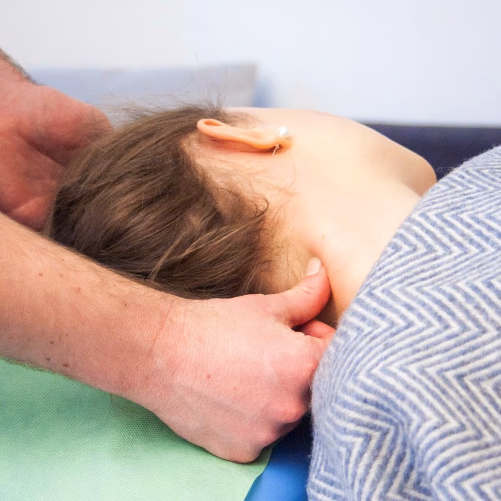 Hände massieren den Nacken einer Frau die unter einer grau gemusterten Decke liegt