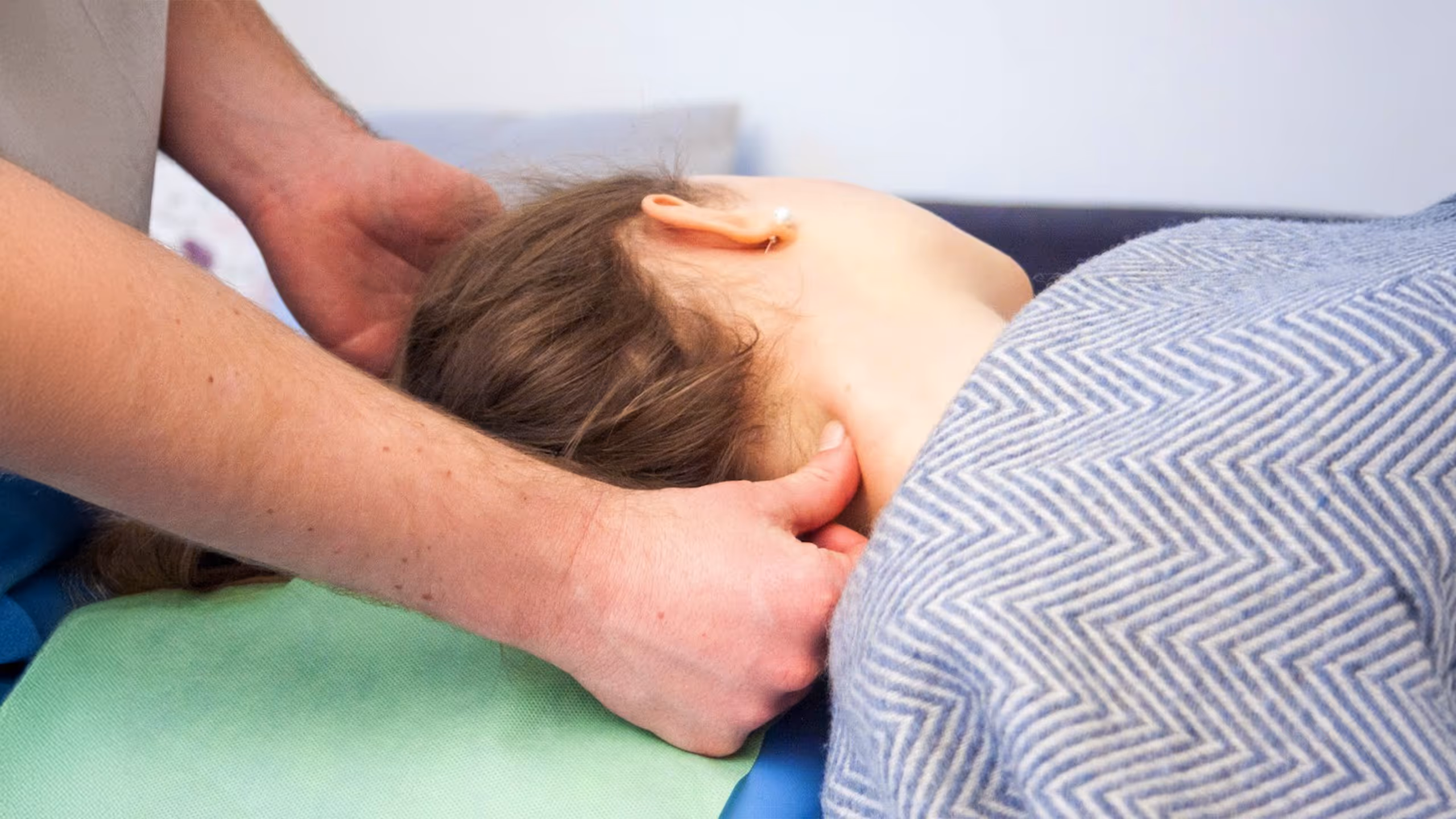 Hände massieren den Nacken einer Frau die unter einer grau gemusterten Decke liegt