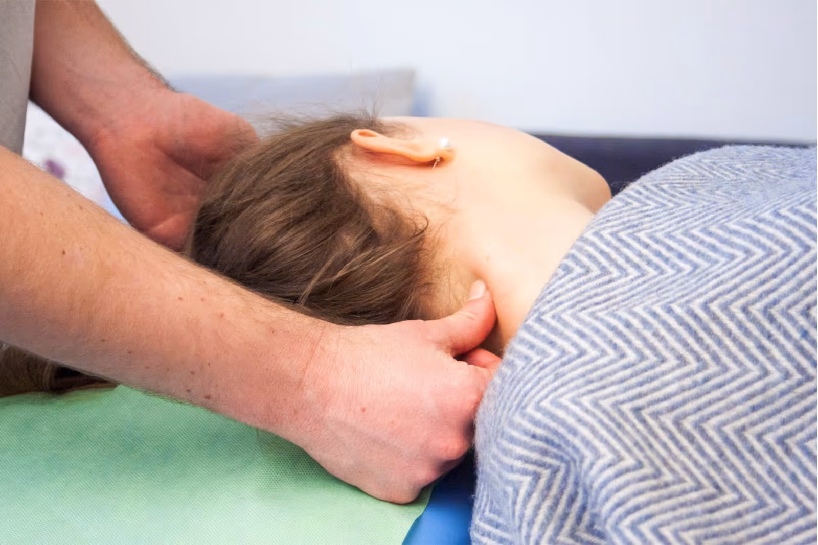 Hände massieren den Nacken einer Frau die unter einer grau gemusterten Decke liegt