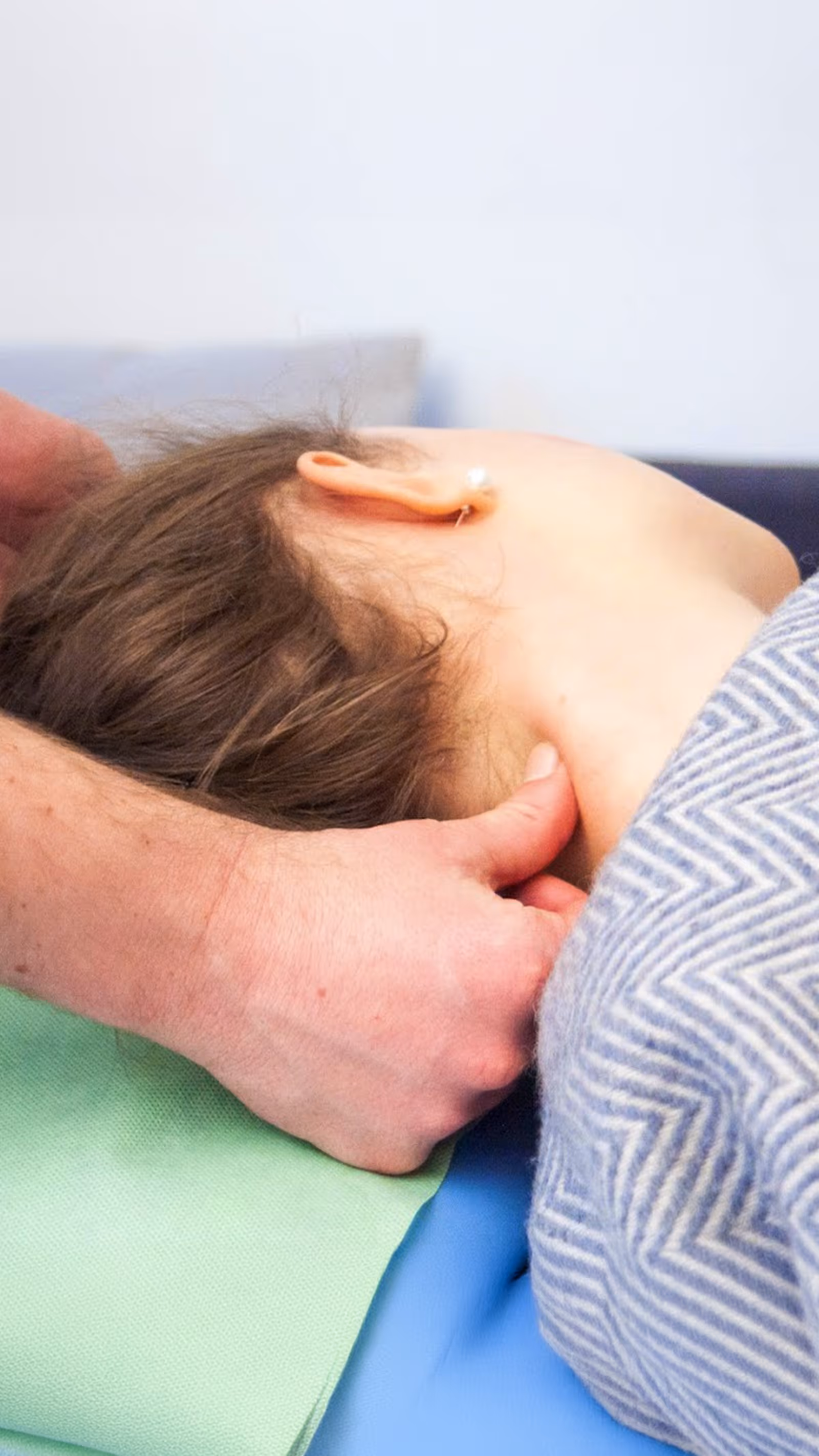 Hände massieren den Nacken einer Frau die unter einer grau gemusterten Decke liegt