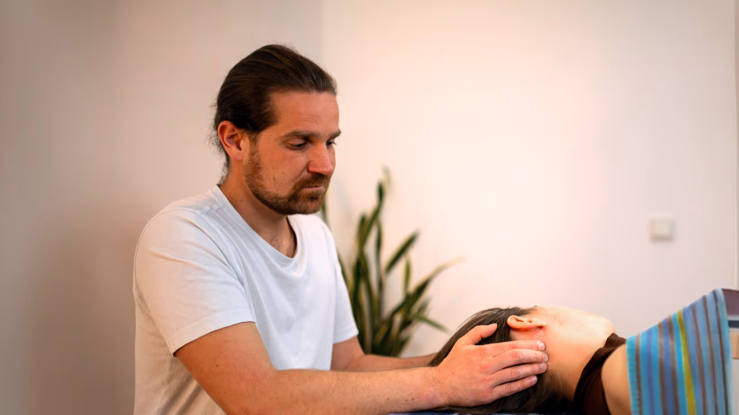 Mann mit zusammengebundenem Haar gibt einer Person eine Kopfmassage die auf einem Massagetisch mit blau karierter Decke liegt