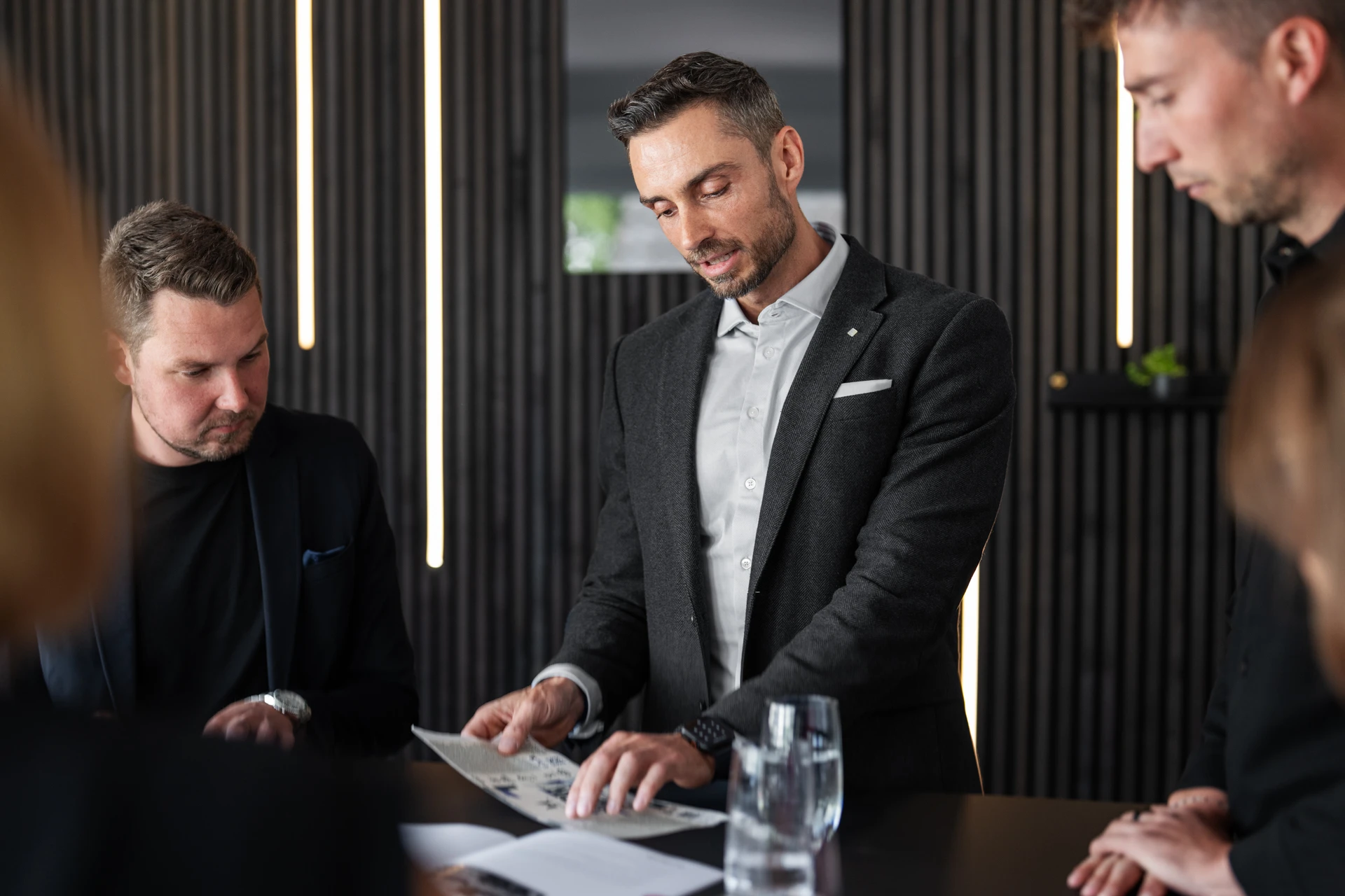 Man in a dark blazer and light shirt showing a document to two other focused men during a meeting in a modern office.