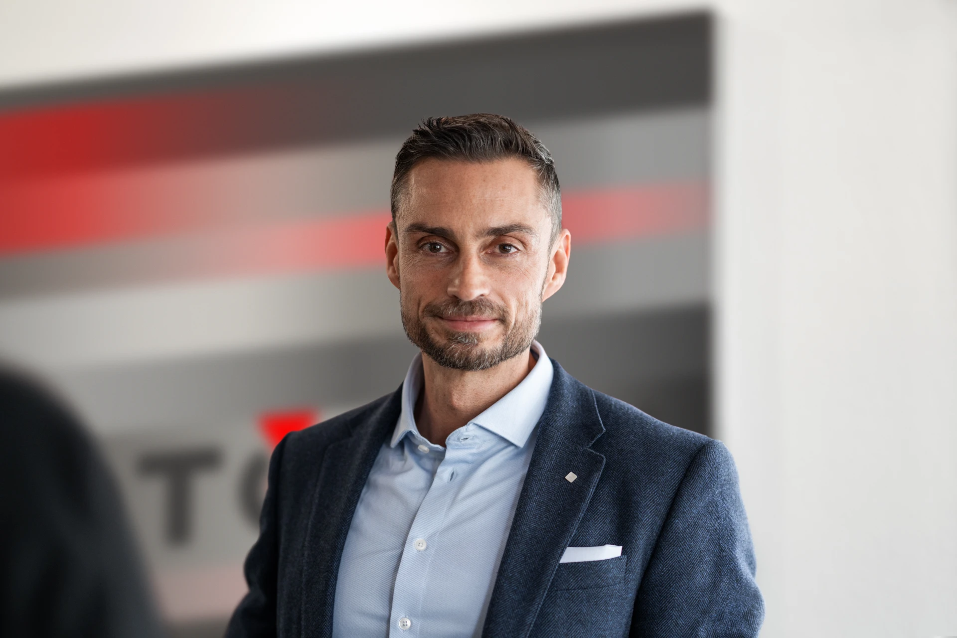 Confident man in a blue blazer and light blue shirt standing indoors with a blurred background featuring red and gray horizontal stripes.