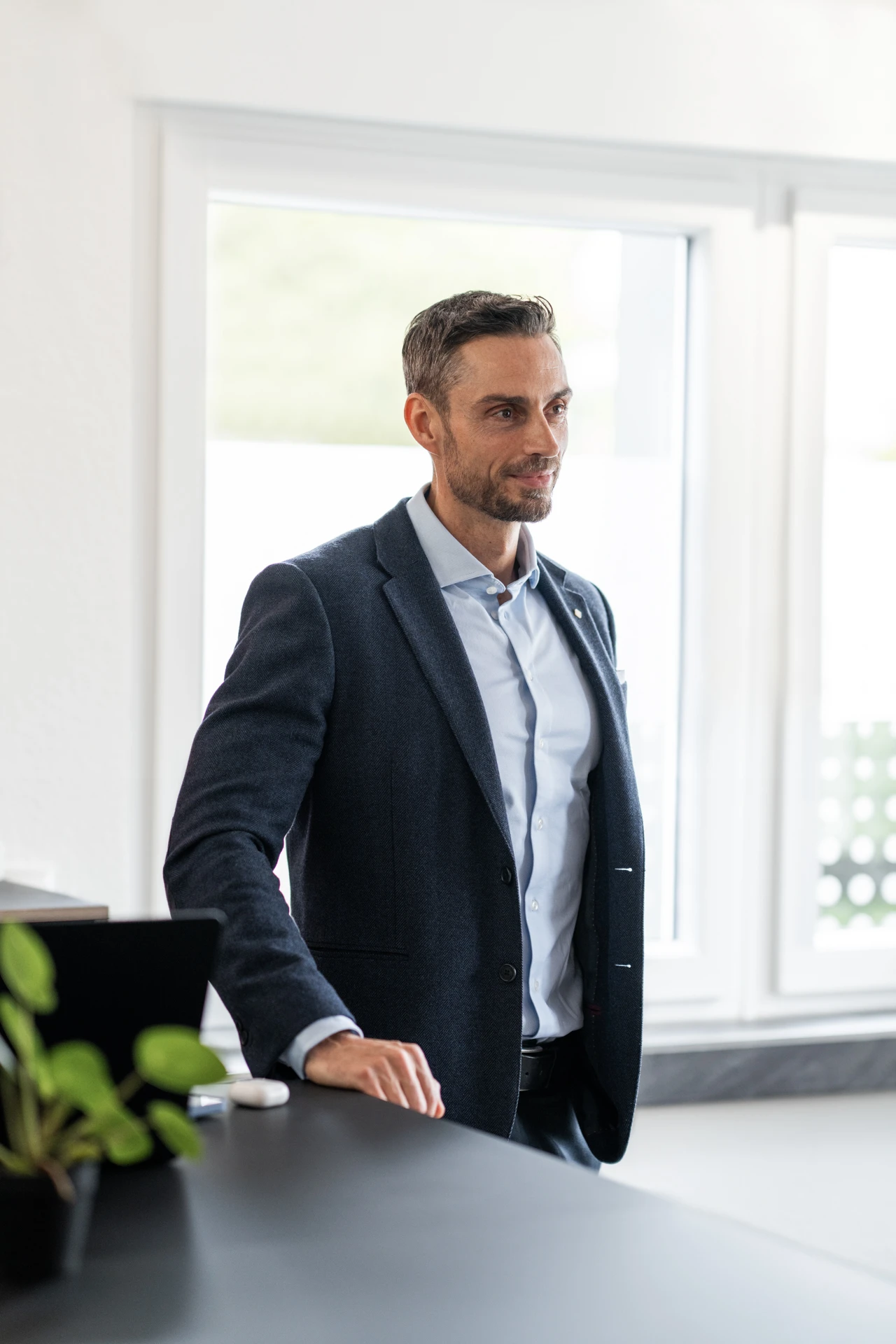 Professional man in a dark suit jacket and light blue shirt standing indoors by a counter near large windows.