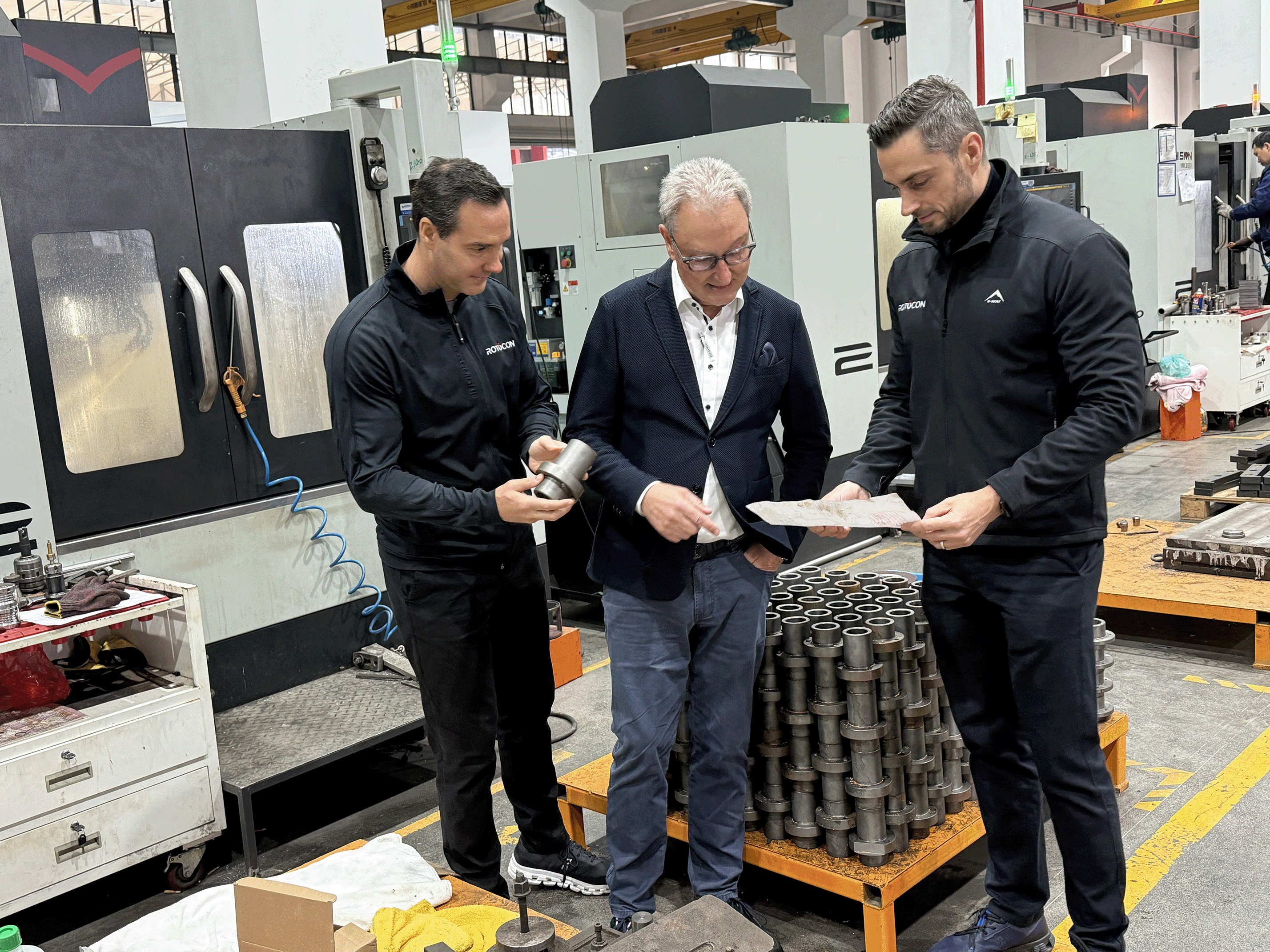 Three men in a manufacturing facility discussing metal parts, one holding a component and another examining a drawing.