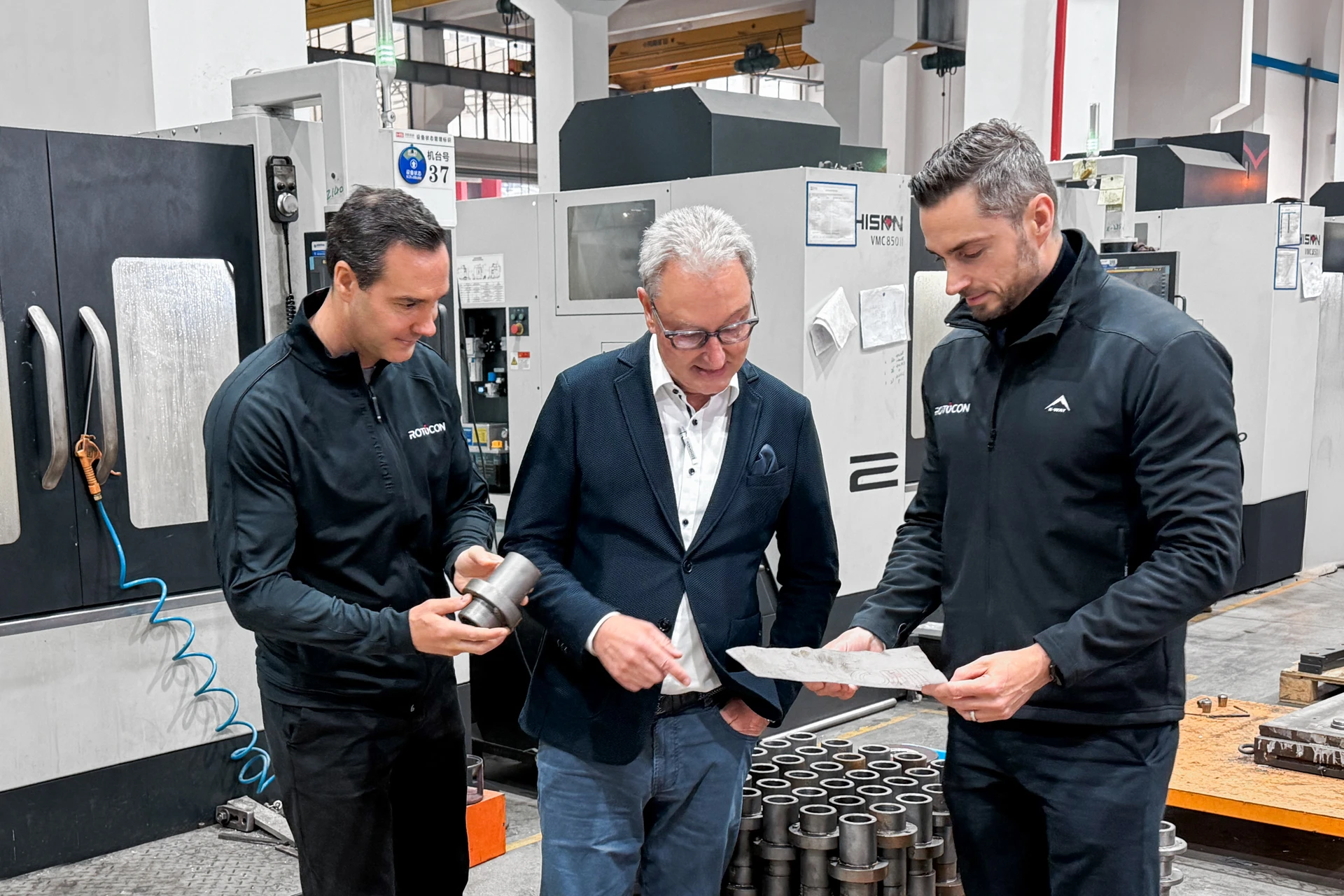 Three men in a manufacturing facility discussing metal parts, one holding a component and another examining a drawing.