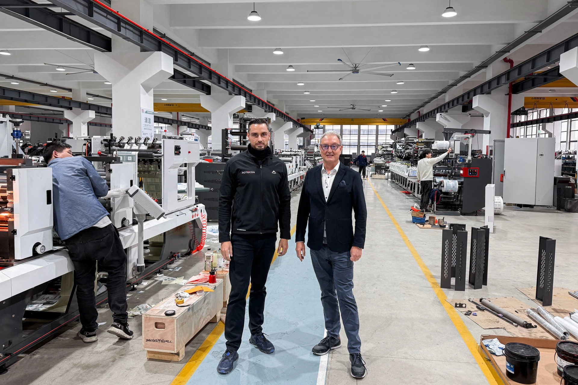 Two men standing in the center aisle of a large, well-lit industrial factory with machinery and workers on both sides.