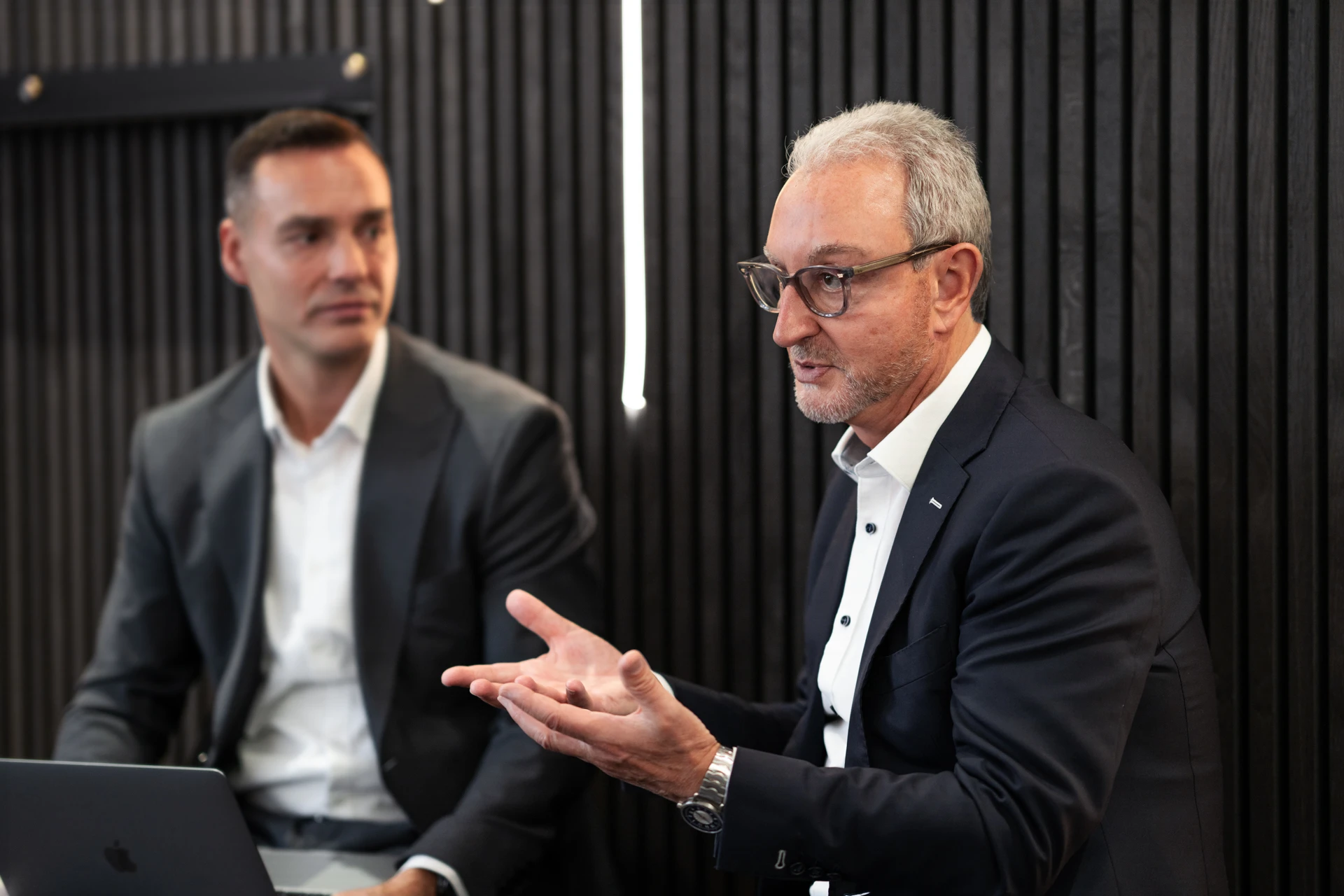 Two men in suits engaged in a conversation, with one gesturing while speaking and the other listening attentively with a laptop.
