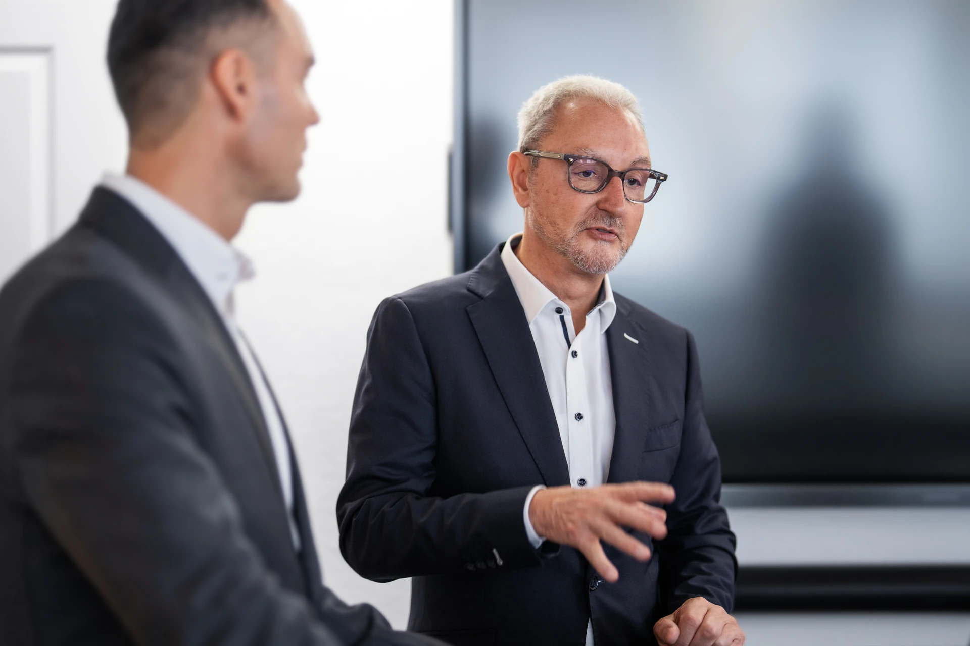 Two men in business suits having a discussion in an office setting with a large screen in the background.