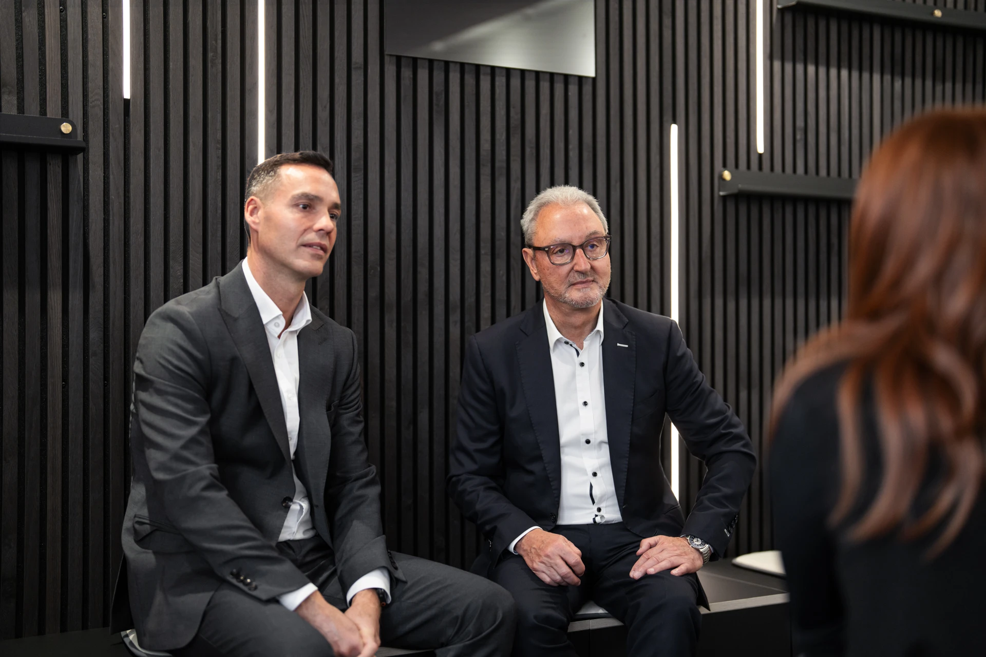 Two businessmen in suits seated and attentively listening to a woman with long hair in a modern office setting with dark vertical panel walls.