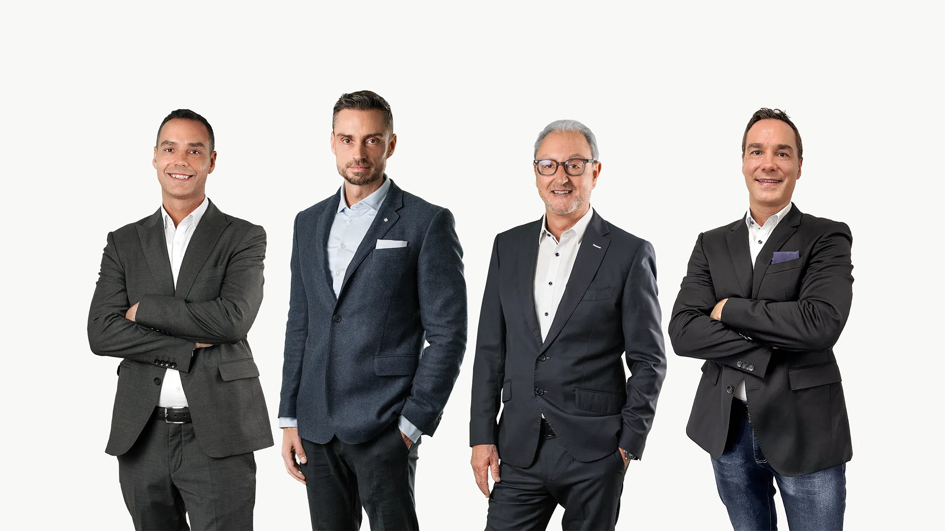 Four men standing side by side against a plain gray background, dressed in business attire with two men crossing their arms and the other two with hands in pockets.