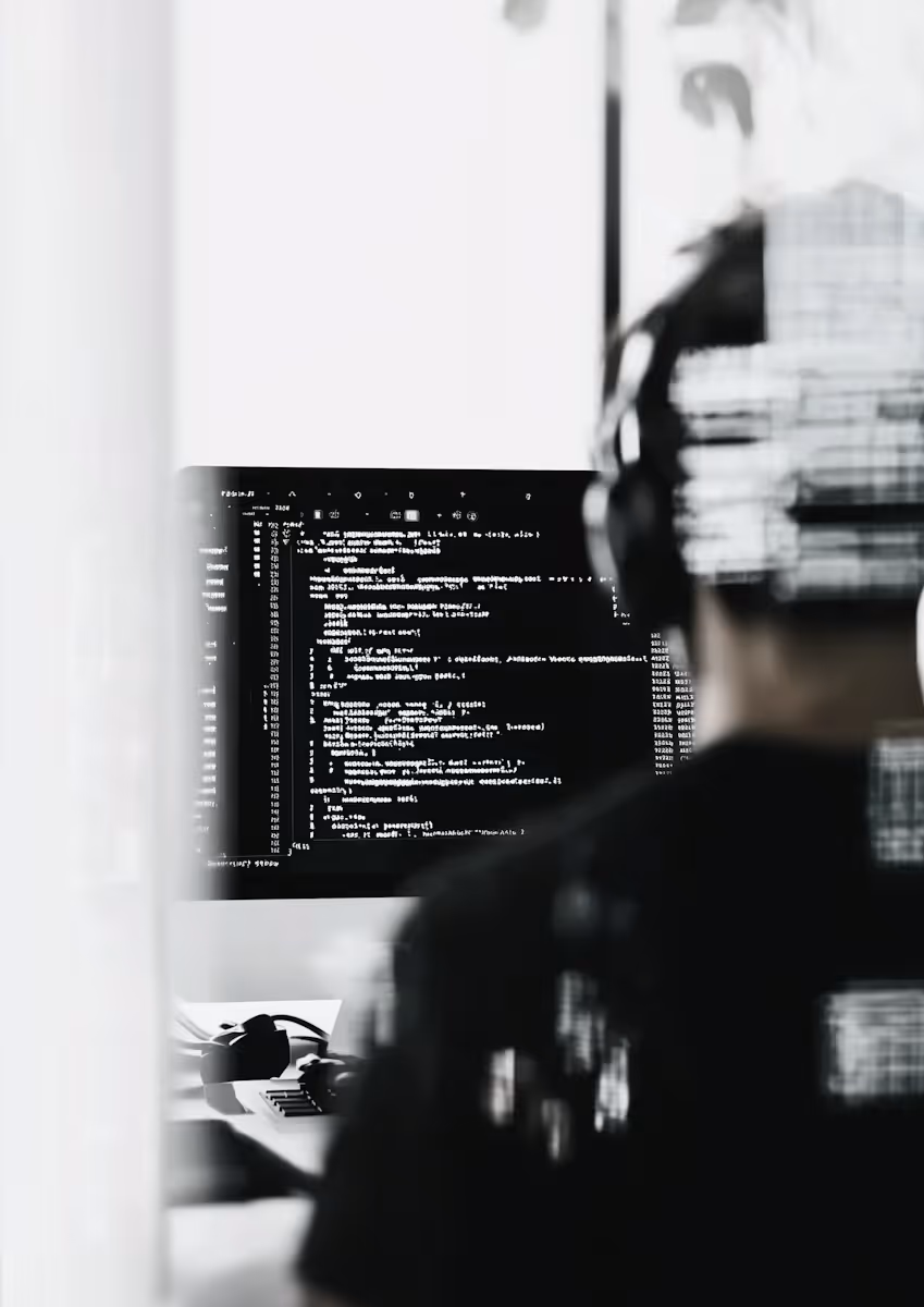 Person med hörlurar som programmerar på en dator med svart skärm och vit kod i ett suddigt, svartvitt motiv.