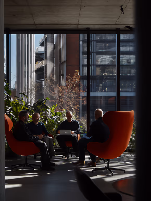 Fyra personer har ett möte i en modern kontorsmiljö med stora fönster och orangea fåtöljer.