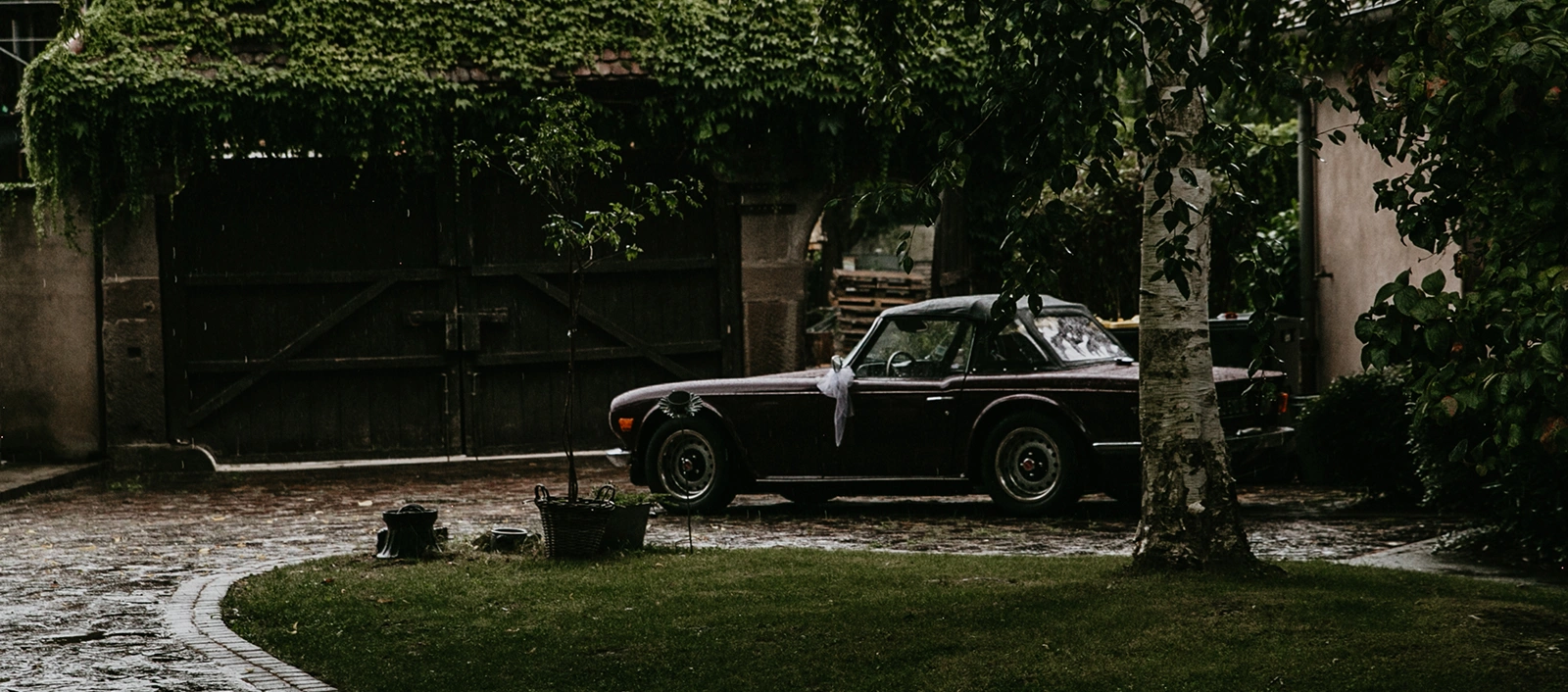Voiture de collection décorée pour un mariage sous une pluie fine devant une porte en bois à La Ferme Marie Hélène.