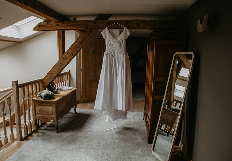 Robe de mariée blanche suspendue dans une chambre élégante de la ferme Marie Hélène avec poutres apparentes et meubles en bois.