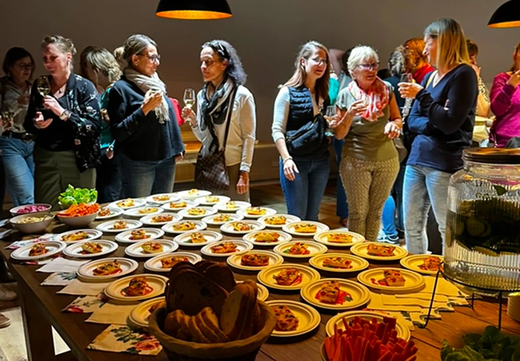 Table remplie d’assiettes de tapas avec un groupe de personnes en arrière-plan, discutant et tenant des verres de vin lors d'un séminaire à la ferme Marie Hélène.