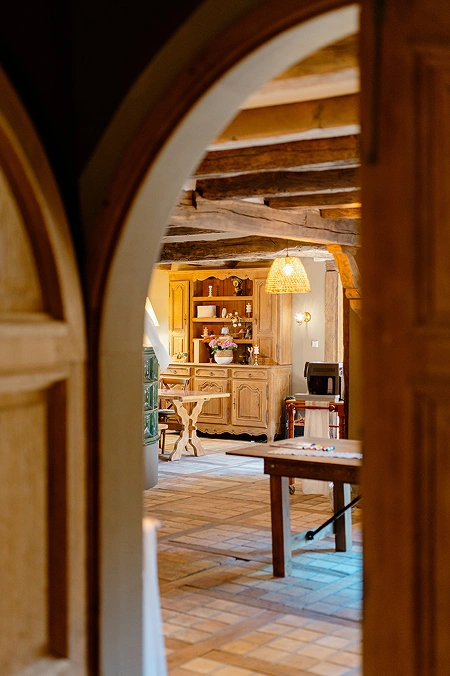 Vue à travers une arche en bois dévoilant une pièce chaleureuse aux poutres apparentes et meubles en bois, typique du cadre authentique de La Ferme Marie Hélène.