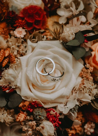 Deux alliances en argent posées sur une rose blanche au centre d’un bouquet de fleurs séchées aux teintes chaleureuses.