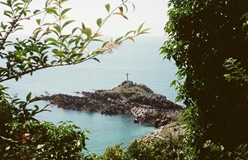 Une petite ile au centre du cadre bordé de branches dans une mer turquoise on y aperçoit une église avec une croix au centre. Photo de Caroline Vincent.