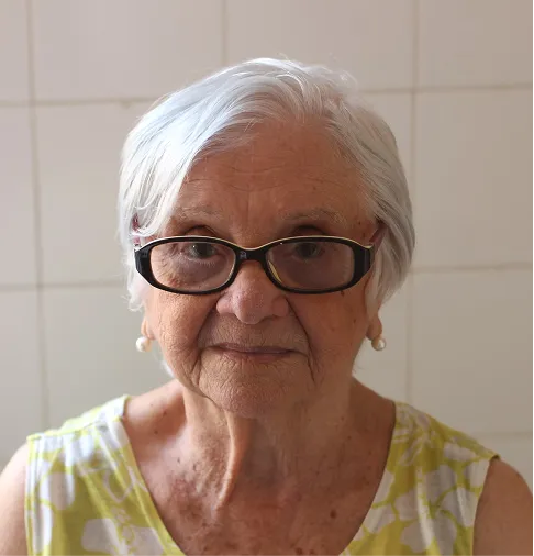 An older woman wearing glasses in front of a tiled wall.