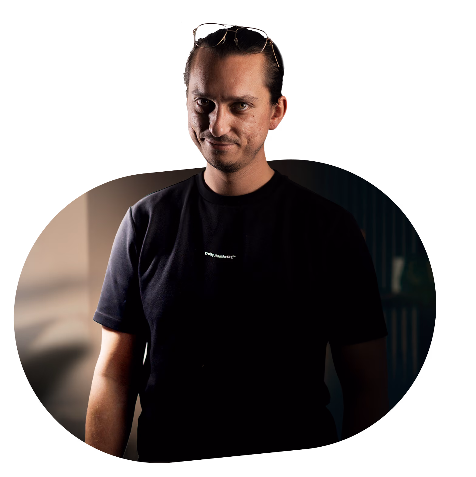 Man with glasses on head wearing a black t-shirt and looking at the camera in dim light.