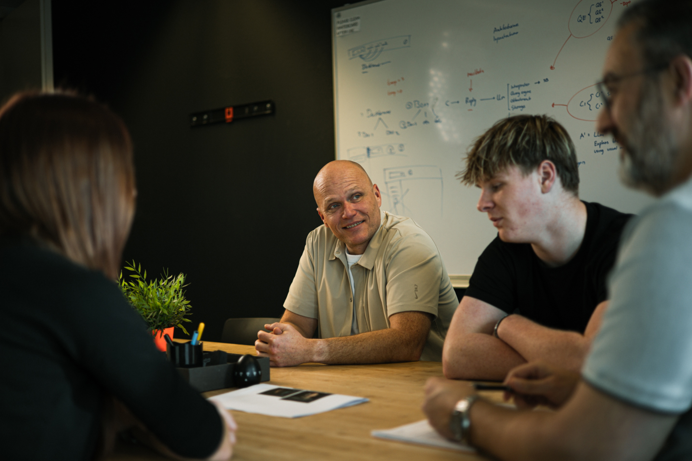 Four people in a meeting room with a whiteboard covered in handwritten notes behind them.